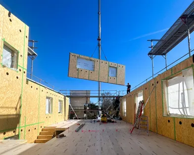Montage einer Holztafelwand mit Fensterausschnitt per Kran auf einer Baustelle eines Holzrahmenbaus bei sonnigem Wetter