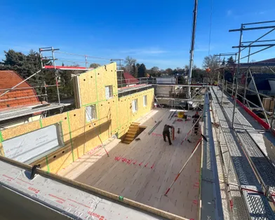 Montage einer Holztafelwand mit Fensterausschnitt per Kran auf einer Baustelle eines Holzrahmenbaus bei sonnigem Wetter