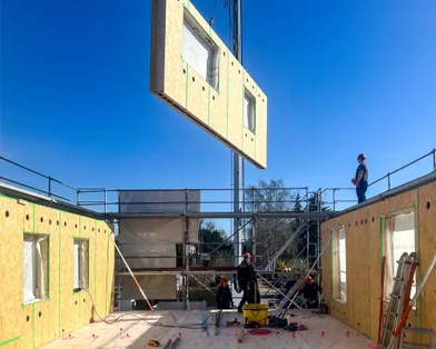 Montage einer Holztafelwand mit Fensterausschnitt per Kran auf einer Baustelle eines Holzrahmenbaus bei sonnigem Wetter